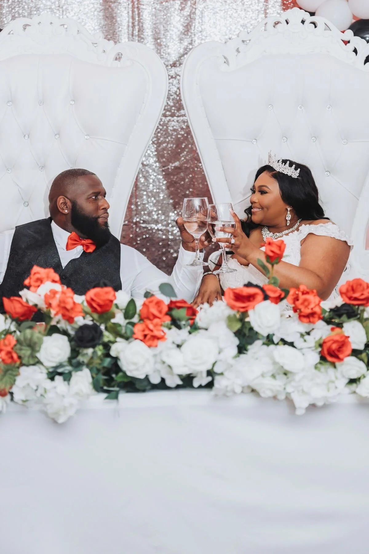 A bride and groom sitting at a wedding table, raising glasses in a toast, with a backdrop of pink, black, and white balloons, and a floral arrangement of red, black, and white flowers in front of them.