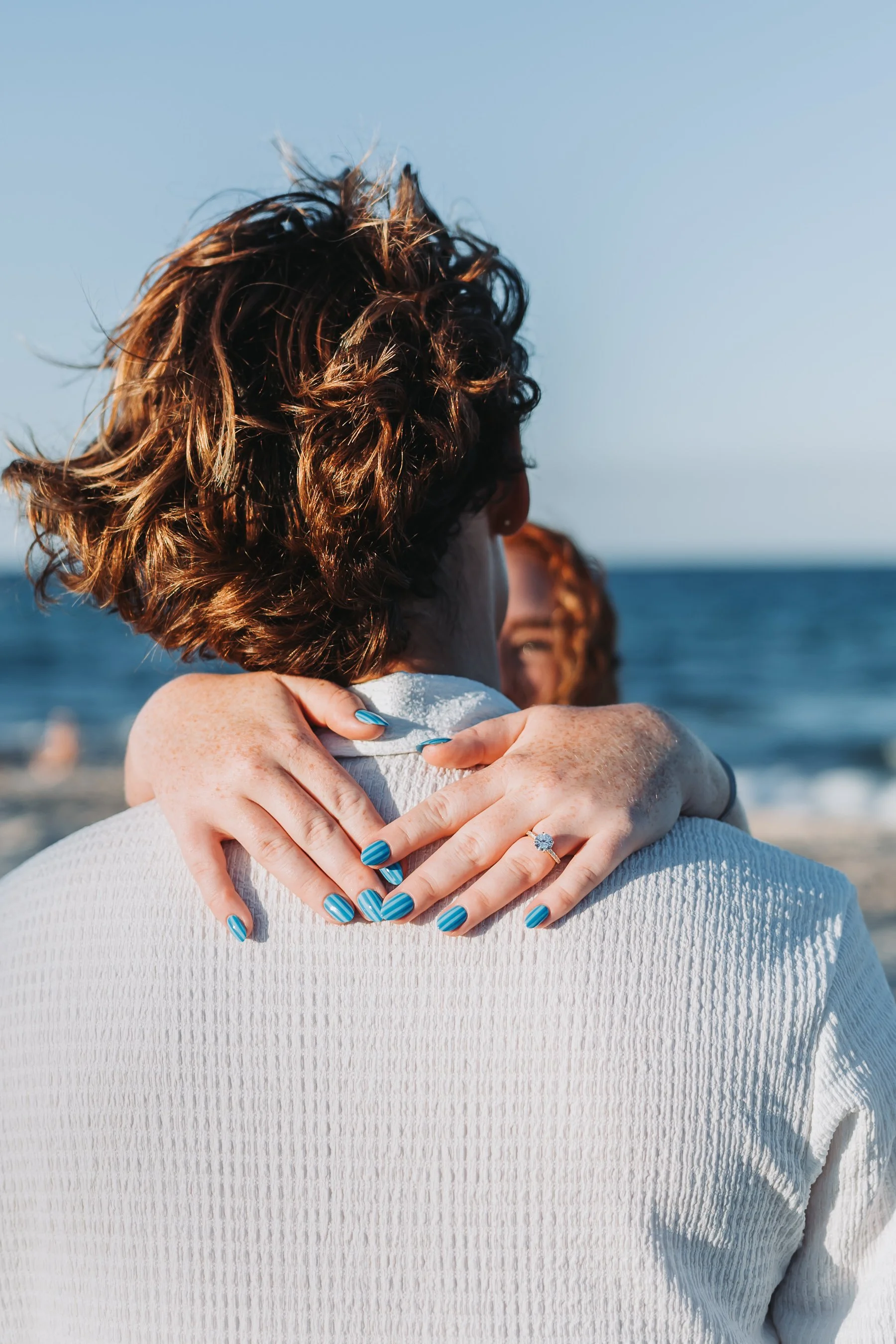 Close-up of a woman with curly hair embracing someone at the beach, with ocean and clear sky in the background.