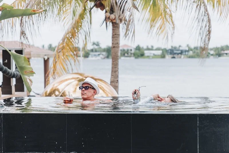 A man wearing sunglasses and a hat relaxing in a pool next to a lake with palm trees and houses in the background.