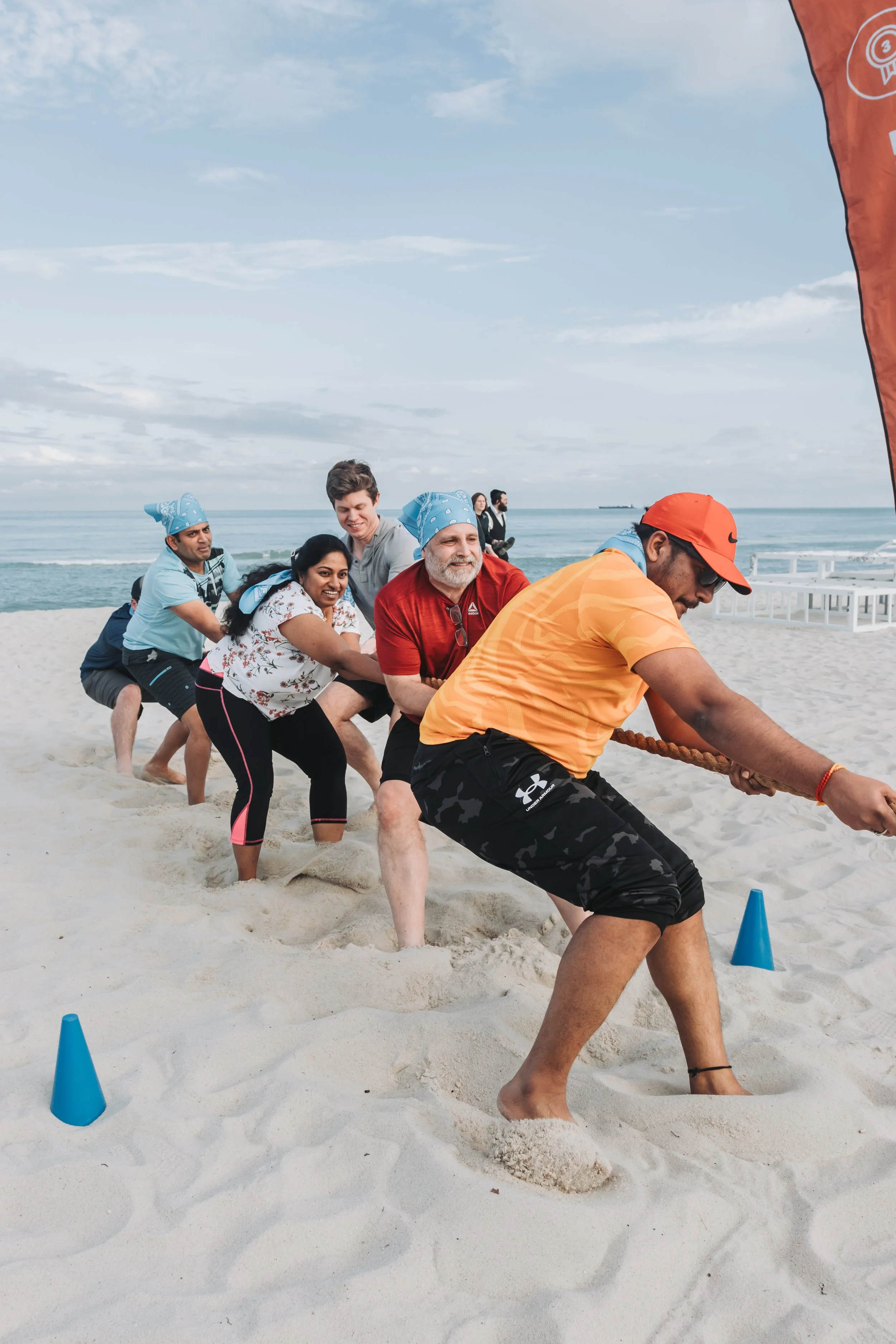 A group of people participating in a tug-of-war game on a sandy beach, with the ocean and a clear sky in the background.