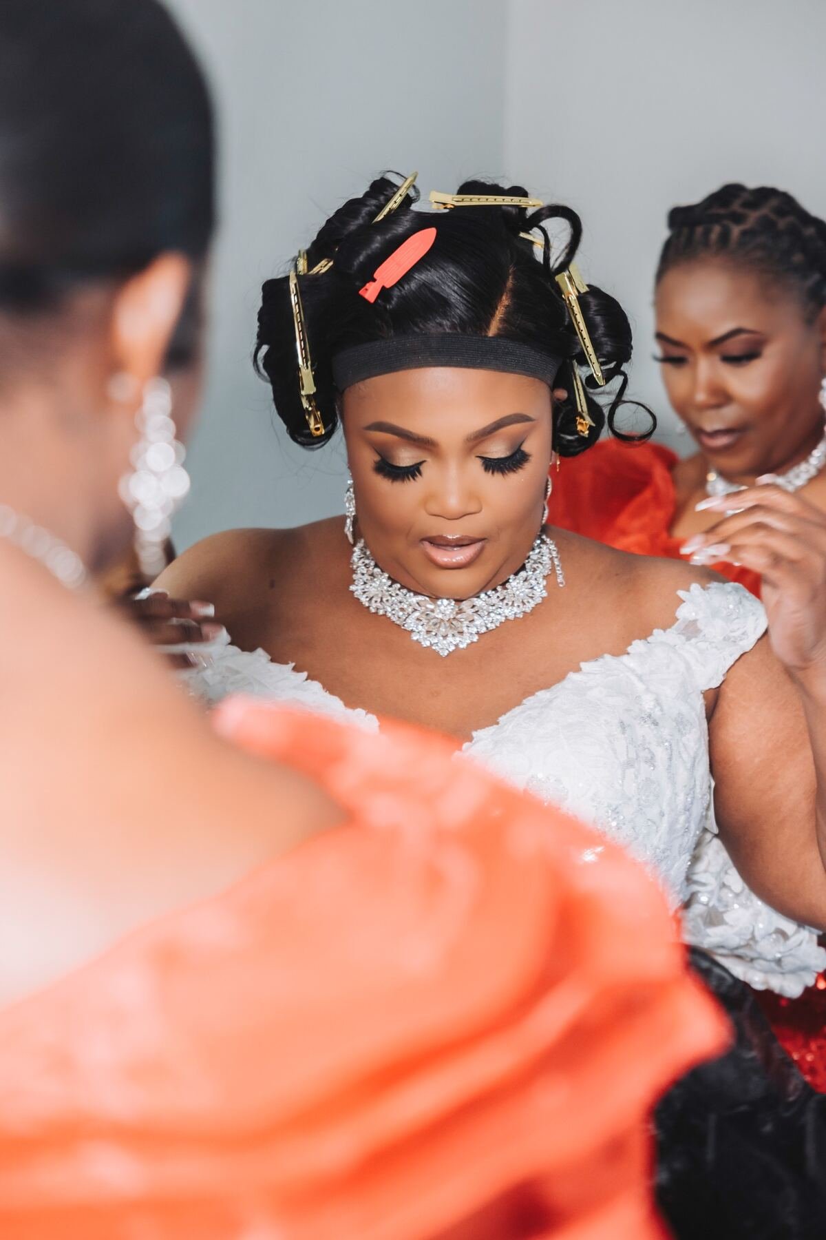 A bride with styled hair in curlers and a black headband, wearing a white lace dress and ornate jewelry, is being assisted by women with jewelry and styled hair.