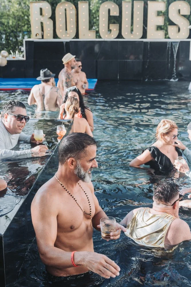 People lounging and socializing in an outdoor pool with a large lit sign reading 'ROLGUES' in the background.