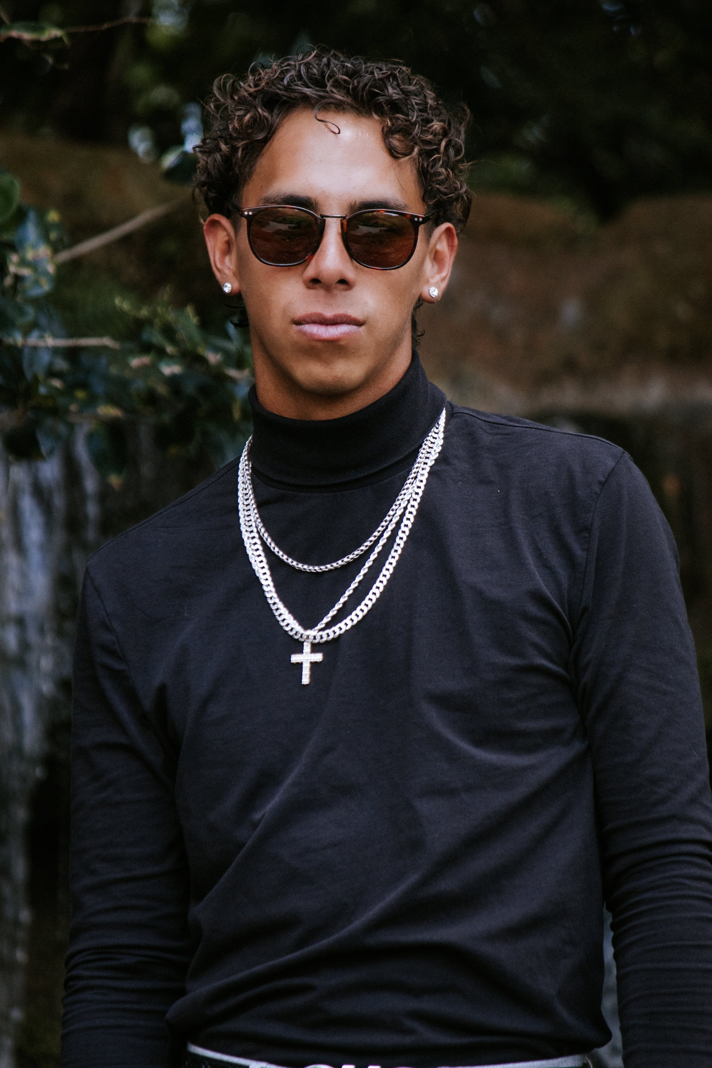 Young man with curly hair wearing sunglasses, earrings, layered chains with a cross pendant, and a black turtleneck in an outdoor setting.