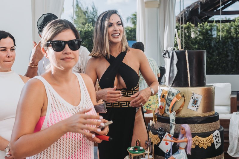 Women at a birthday celebration with a black and gold cake decorated with playing cards and a doll, outdoors with greenery in the background.