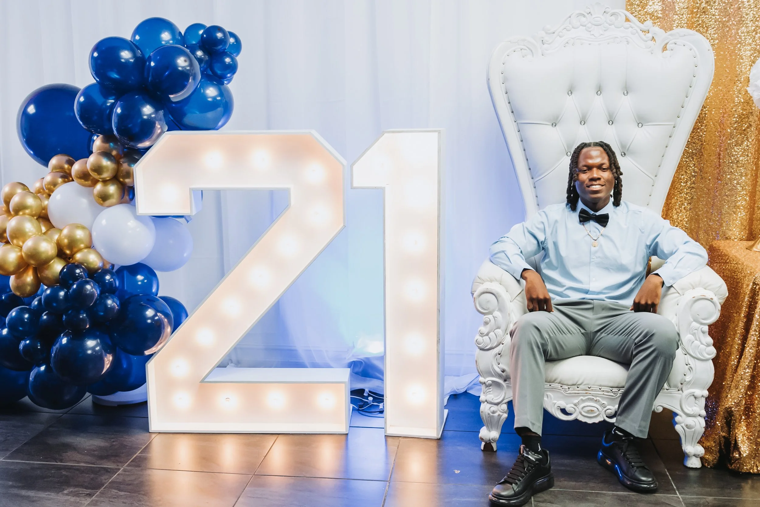 Man sitting on a white ornate throne at a celebration event, with a large illuminated number 21 and balloon decorations nearby.