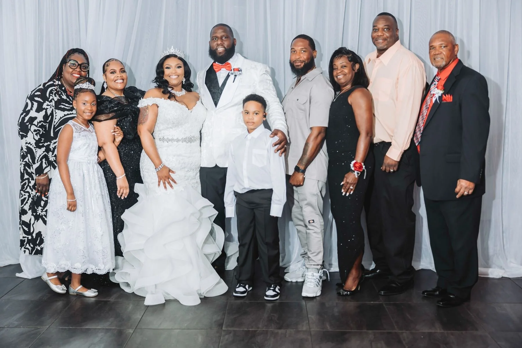 Group of 11 people posing in front of a white curtain, including a bride and groom, children, and family members at a wedding celebration.
