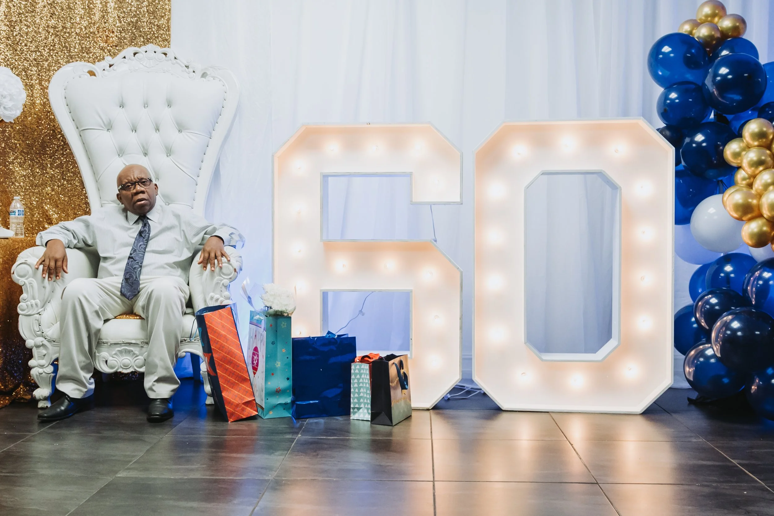 A man in formal attire sitting on an ornate white throne-like chair at a celebration or party with a backdrop of gold, blue, and white decorations, including balloons and gift bags.