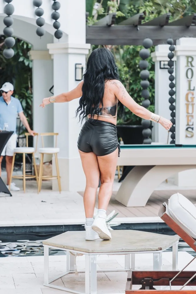 Woman with long black hair in a black leather top and shorts dancing on a small table near a pool with poolside furniture and a life-sized pool table in the background.
