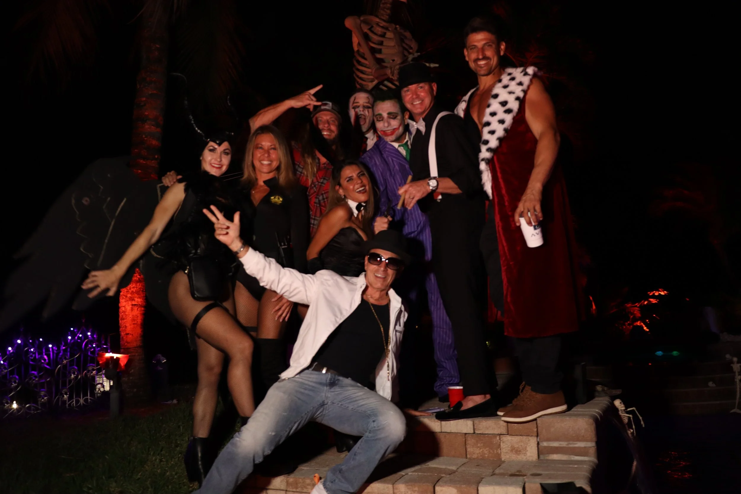 Group of people dressed in costumes, some with clown makeup, posing outdoors at night with skeleton decoration and colorful lights in the background.