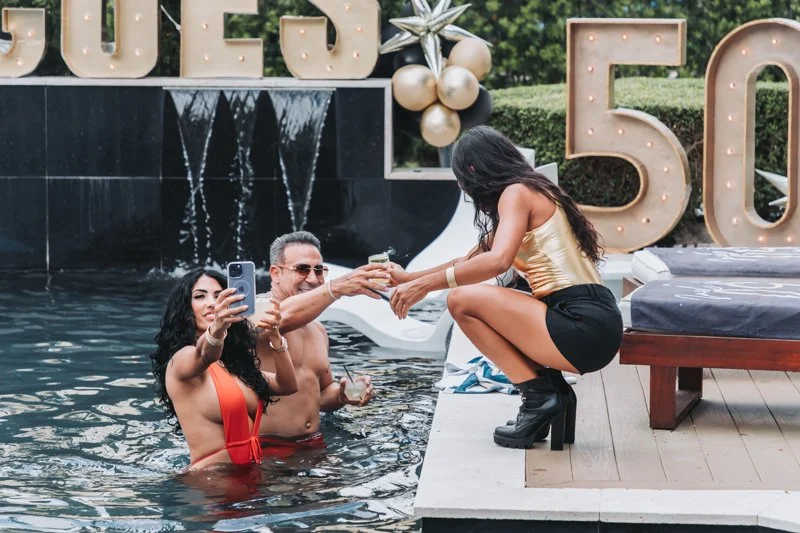 Three people at a poolside celebration, with large illuminated letters reading 'LOVE 50' in the background, and gold and white balloon decorations.