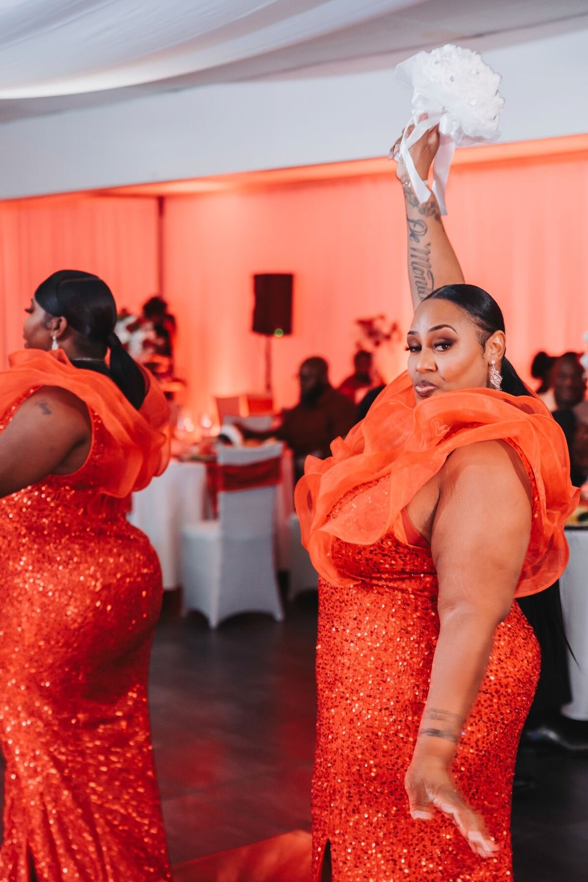 Woman in an orange sequined gown with sheer orange ruffled details, holding a white bouquet above her head, at a formal event with orange lighting and other guests in the background.