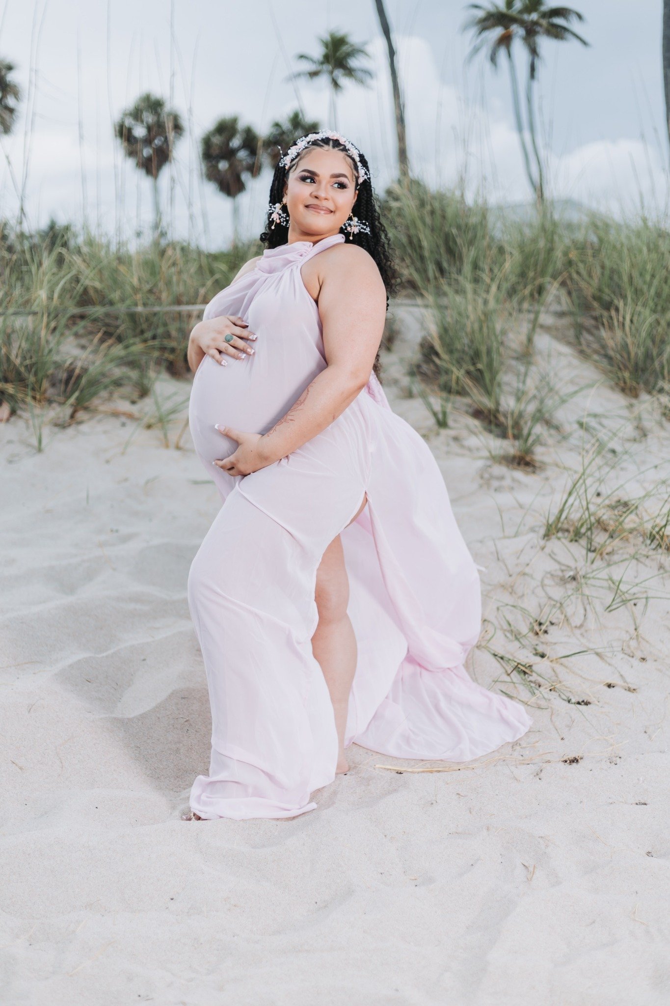 A pregnant woman in a flowing pink dress stands on a sandy beach with tall grass and palm trees in the background, smiling and gently holding her belly.