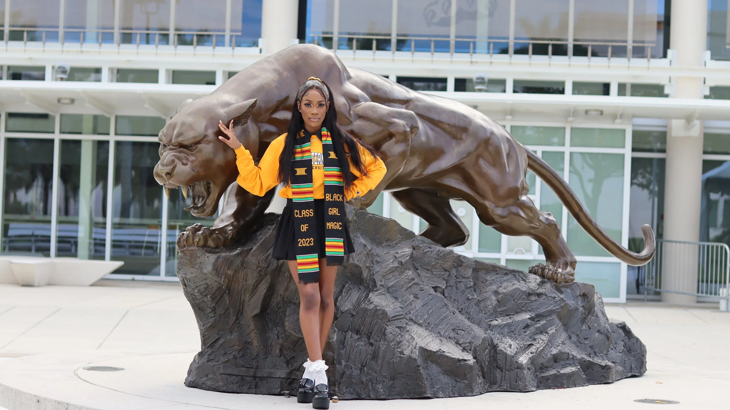 A young woman standing in front of a large lion statue with her hand resting on its head. She is wearing a yellow sweatshirt, a black skirt, and a colorful graduation stole that reads 'Black Girl Magic 2023'. She has long dark hair and is outdoors in