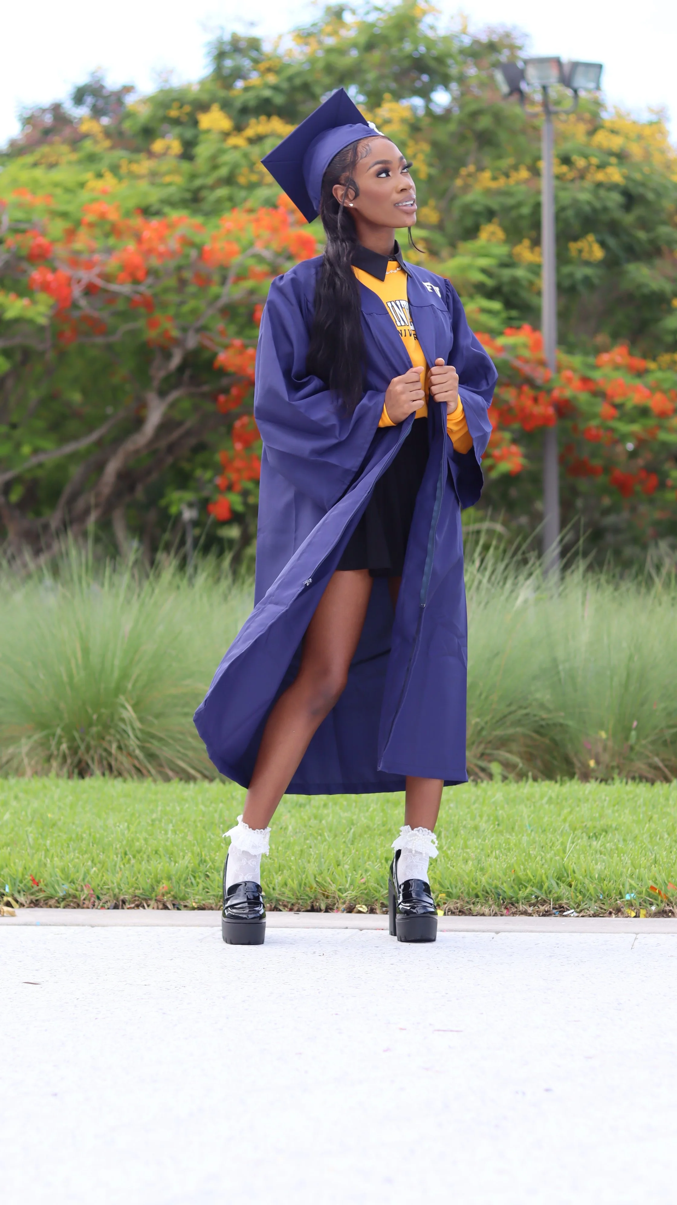 A young woman in a blue graduation gown and cap standing outdoors on grass with colorful trees in the background, wearing platform shoes and white socks.