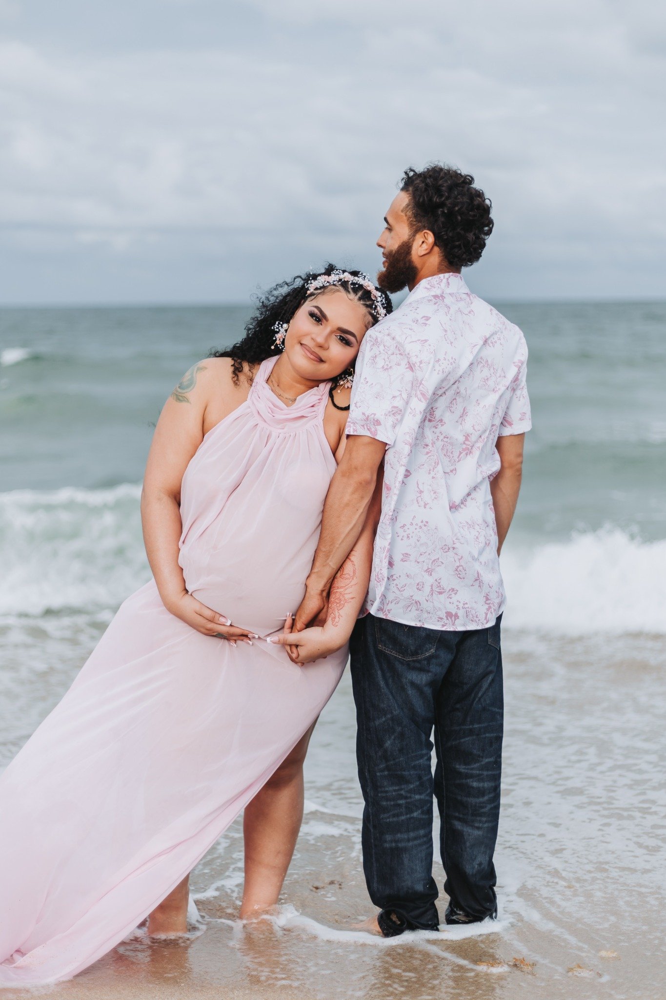 A pregnant woman and a man stand at the shoreline, with the woman resting her head on the man's shoulder. They are dressed in light, summery clothes and are barefoot in the sand, with ocean waves and a cloudy sky in the background.