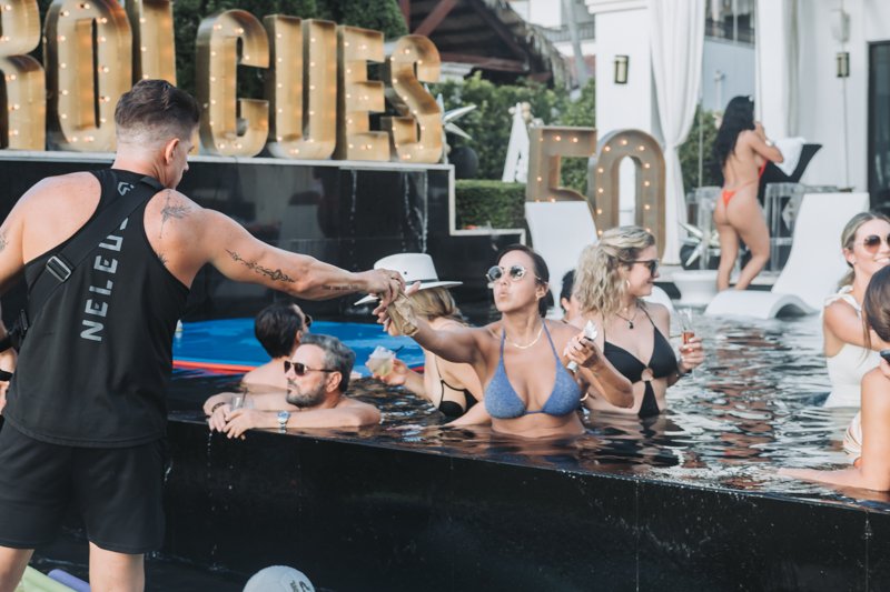 People enjoying a pool party at a nightclub, with a man handing out drinks to women in the pool, and large illuminated signs reading '50' and 'LUCE' in the background.