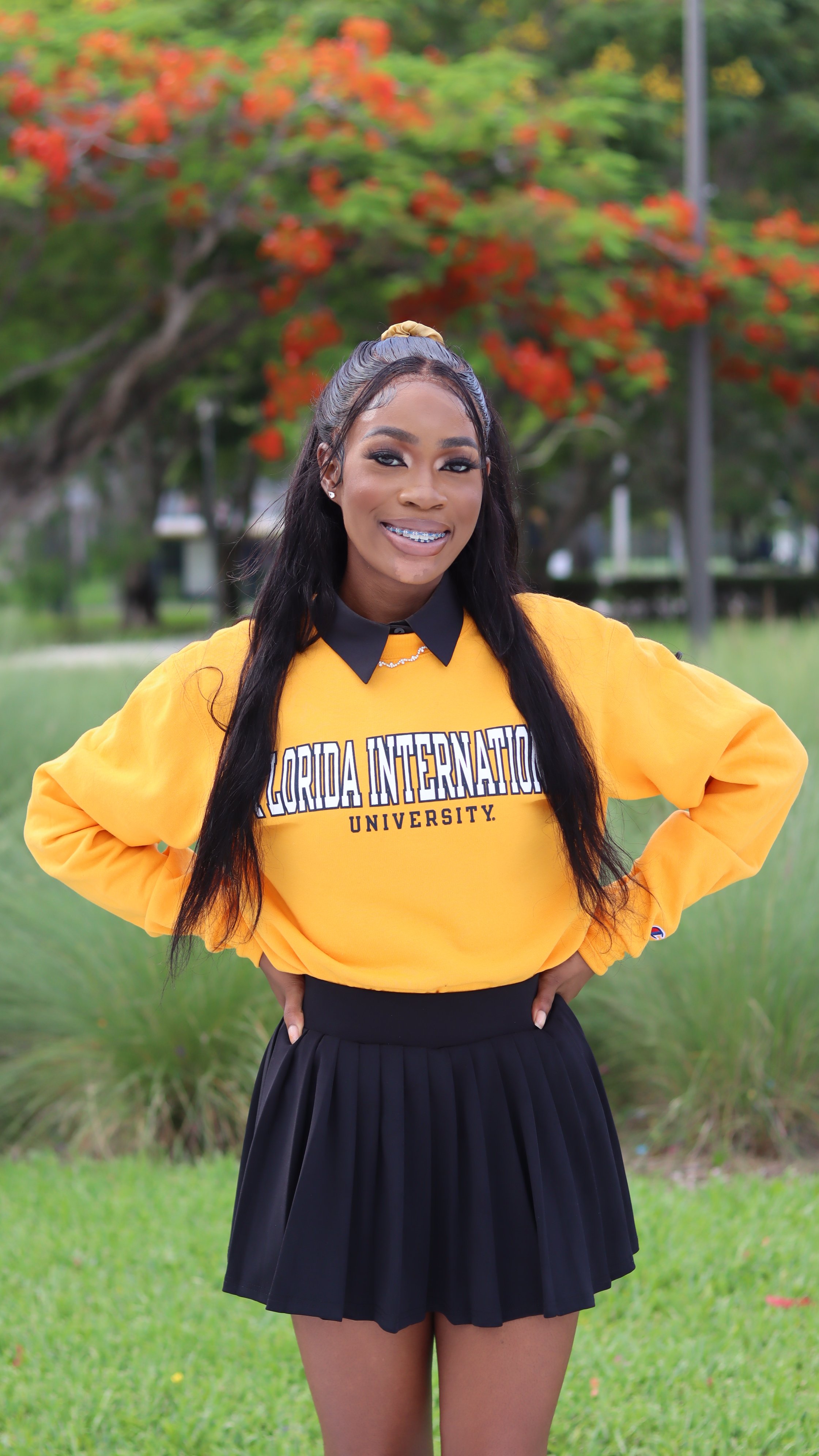 A young woman with long dark hair wearing a yellow 'Florida International University' sweatshirt and a black pleated skirt, standing outdoors in front of trees with orange flowers, smiling with braces.
