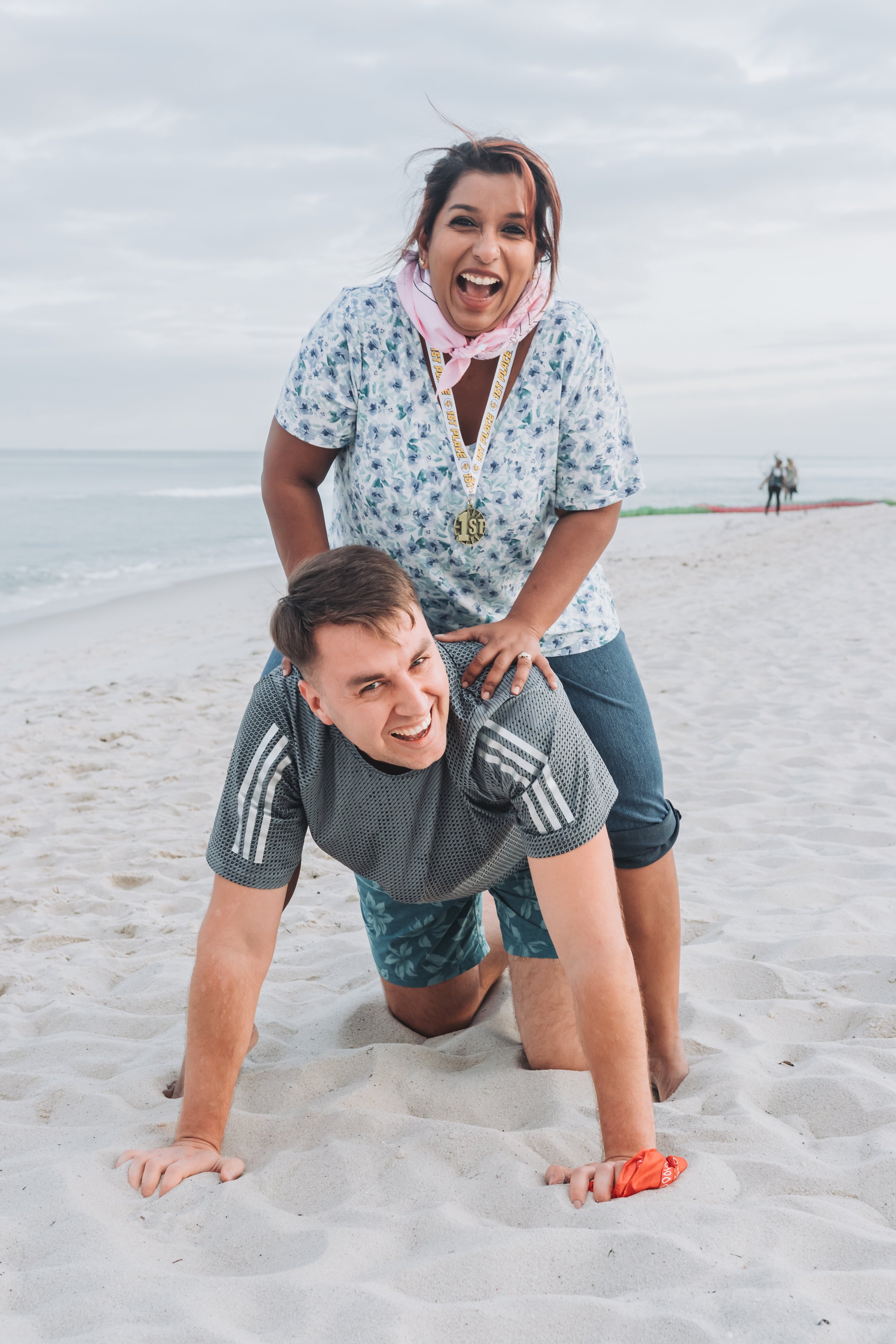 Two people forming a human pyramid on a sandy beach with ocean and cloudy sky in the background. The person on the bottom is kneeling and smiling, while the person on top is standing and laughing, wearing a medal and a pink scarf.