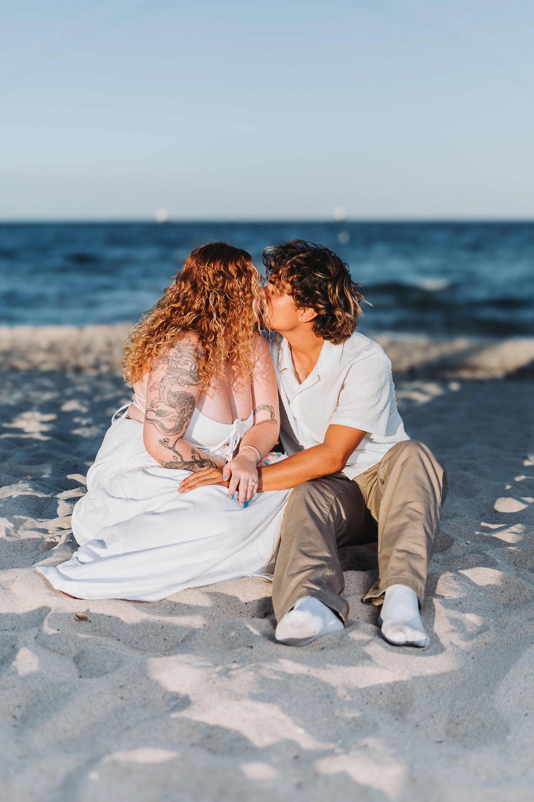 A couple sitting on the sandy beach, kissing with the ocean and blue sky in the background.