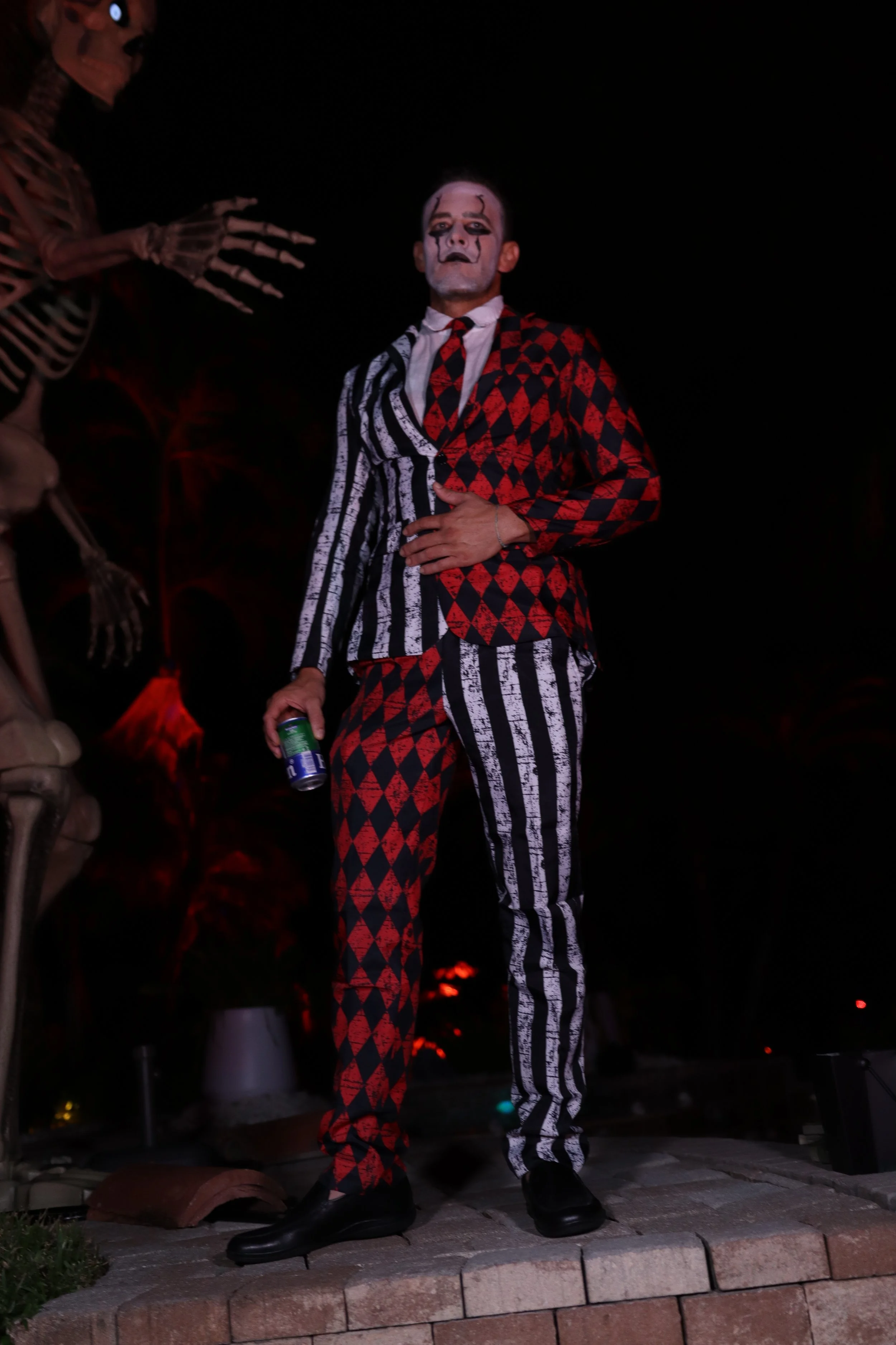 A person dressed as a clown with white face paint, black makeup around the eyes, wearing a red and black patterned suit and a black and white striped tie, holding a soda can, standing on a brick sidewalk at night with skeleton props nearby.