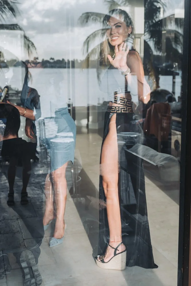 Reflected image of a smiling woman waving, dressed in fashionable clothing, seen through a glass window with reflections of palm trees and sky outside.