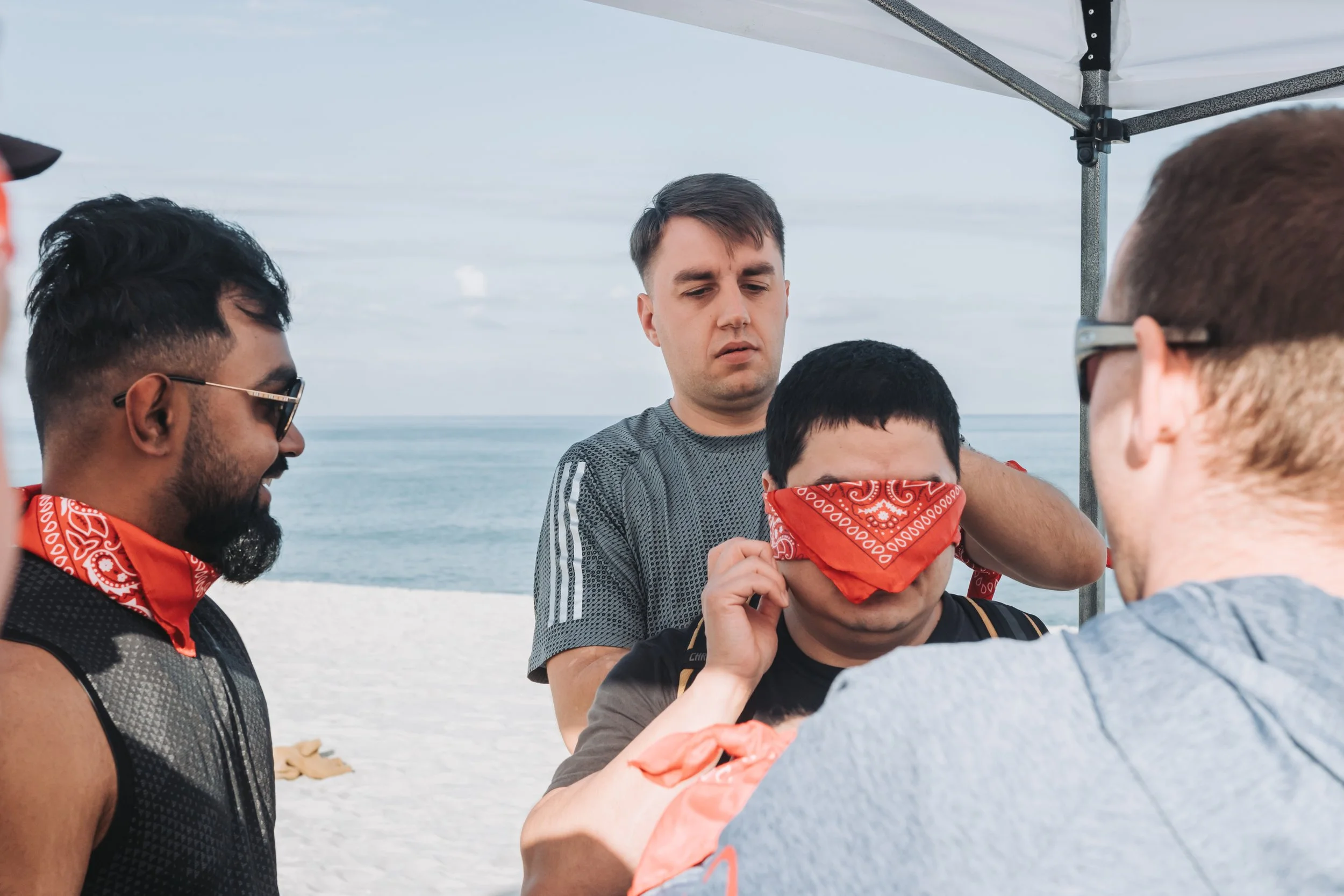 Group of young men standing on the beach, with one man wrapping a red bandana around another man's eyes for a game or activity, with the ocean in the background.