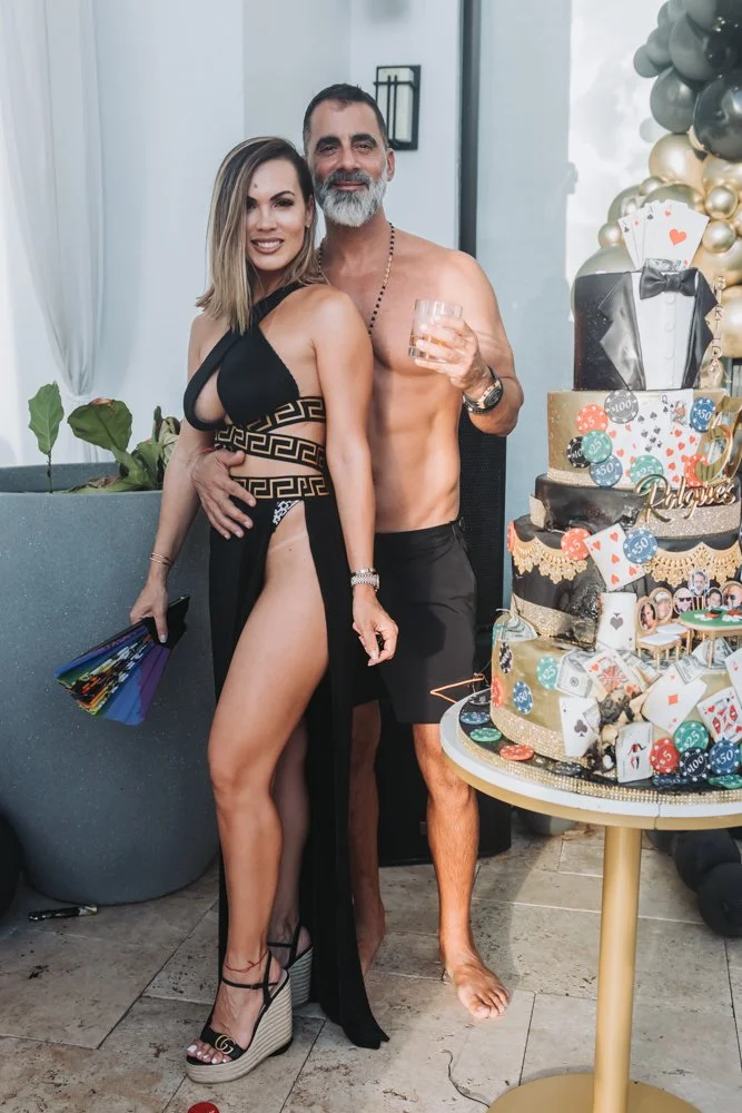 A man and woman posing together at a celebration, with a decorated cake nearby. The woman is wearing a revealing black dress, and the man is shirtless holding a drink. They are smiling at the camera.