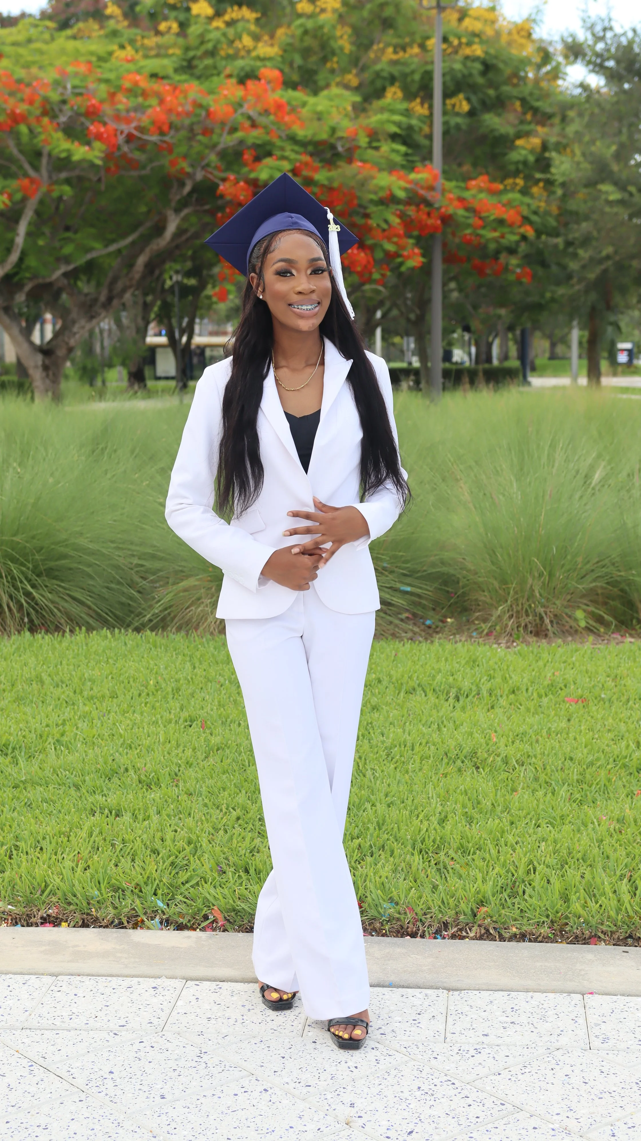 A woman in white graduation attire, including a cap and gown, standing outdoors on a sunny day with green grass and trees with red and yellow leaves in the background, smiling at the camera.