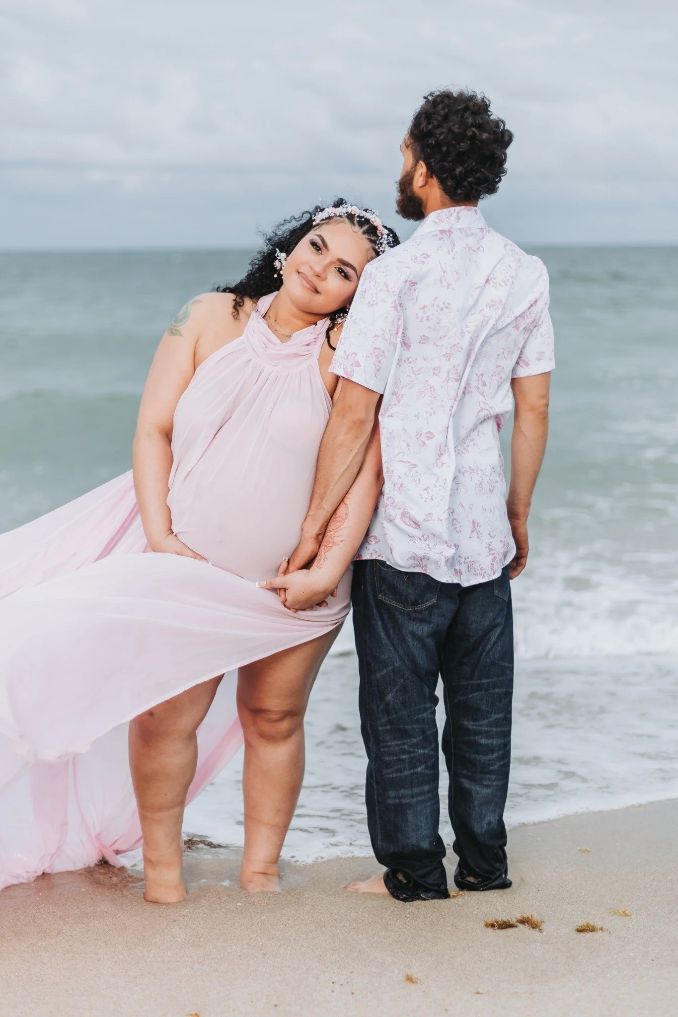 A pregnant woman and a man are standing on the beach with their feet in the sand, facing the ocean. The woman is leaning her head on the man's shoulder, smiling softly, dressed in a flowing pink dress, while the man is wearing a white floral shirt an