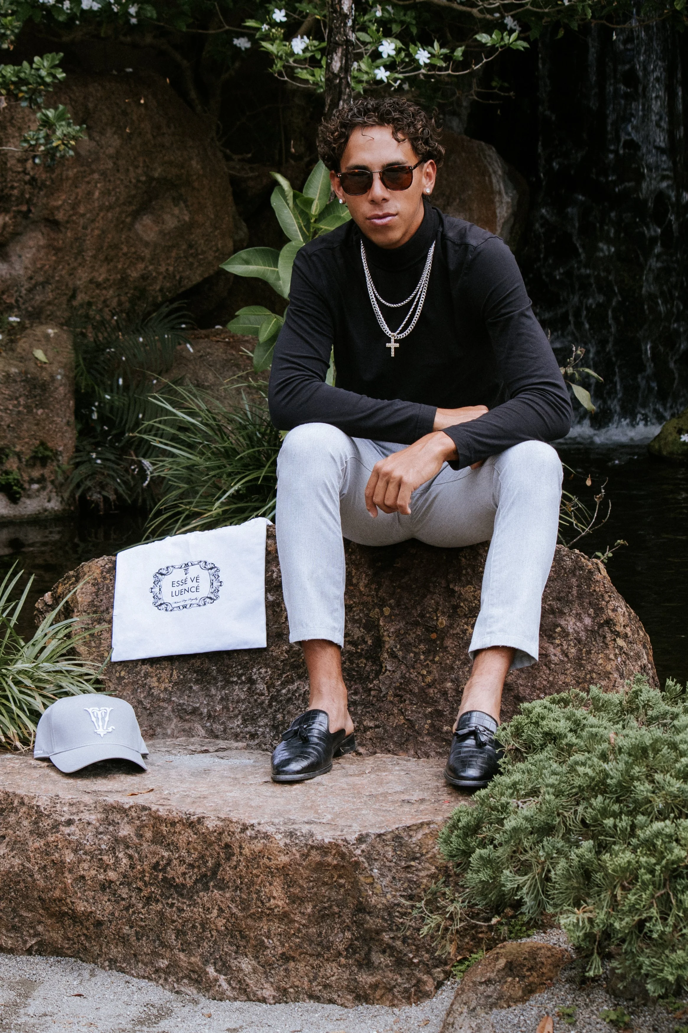 A young man with curly hair and sunglasses is sitting on a large rock in a natural setting with plants and a water feature behind him. He's wearing a black turtleneck, light-colored pants, and black shoes, with layered silver chains and a cross neckl