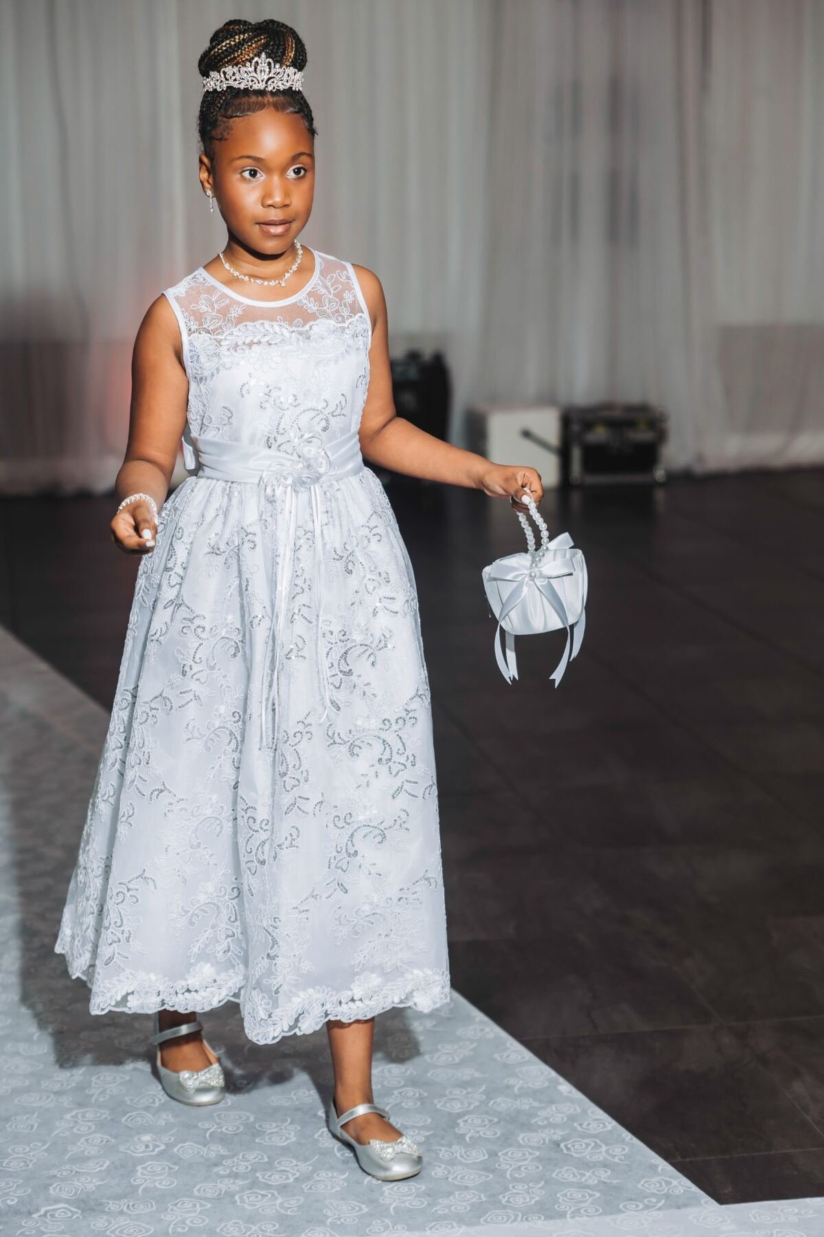 Young girl in a white lace dress, wearing a tiara and pearl jewelry, holding a decorated pillow, walking on a white floral carpet in an event hall.