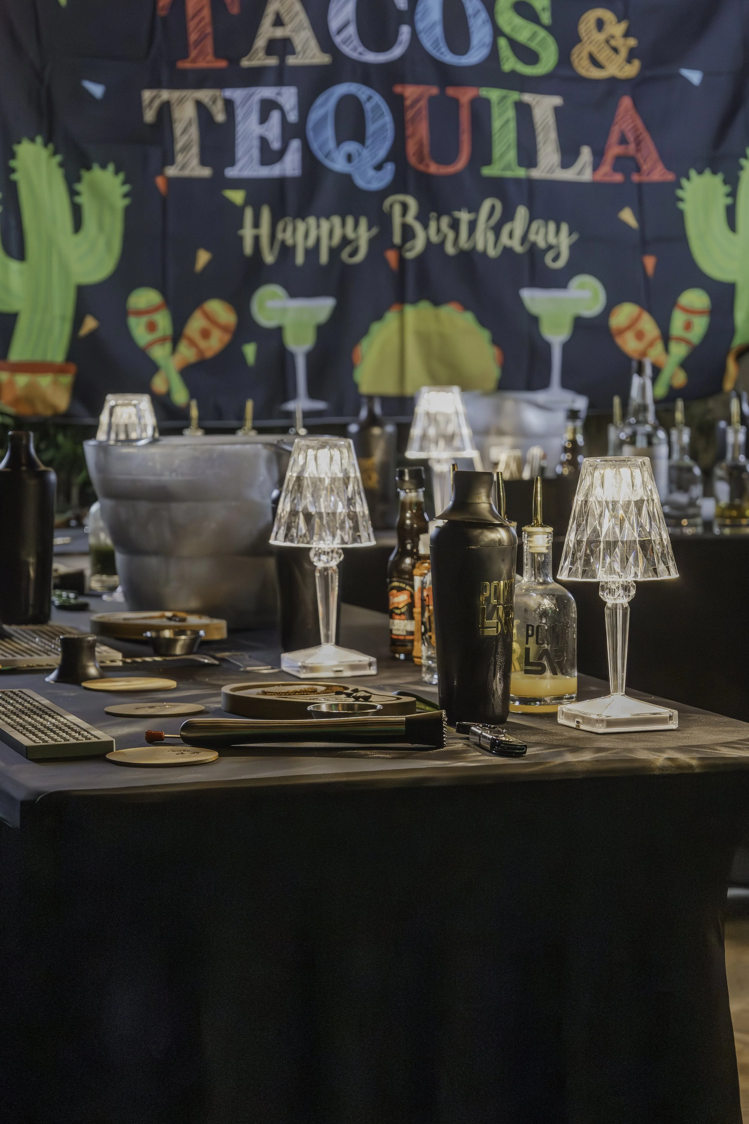 A bar or restaurant counter with drinking glasses, bottles, cocktail shakers, and bar tools in front of a festive birthday banner that reads 'Tacos & Tequila Happy Birthday,' decorated with cactus images.