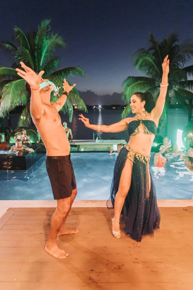 Tropical night scene at a poolside party with dancers in traditional belly dance costumes, palm trees, and a body of water in the background.