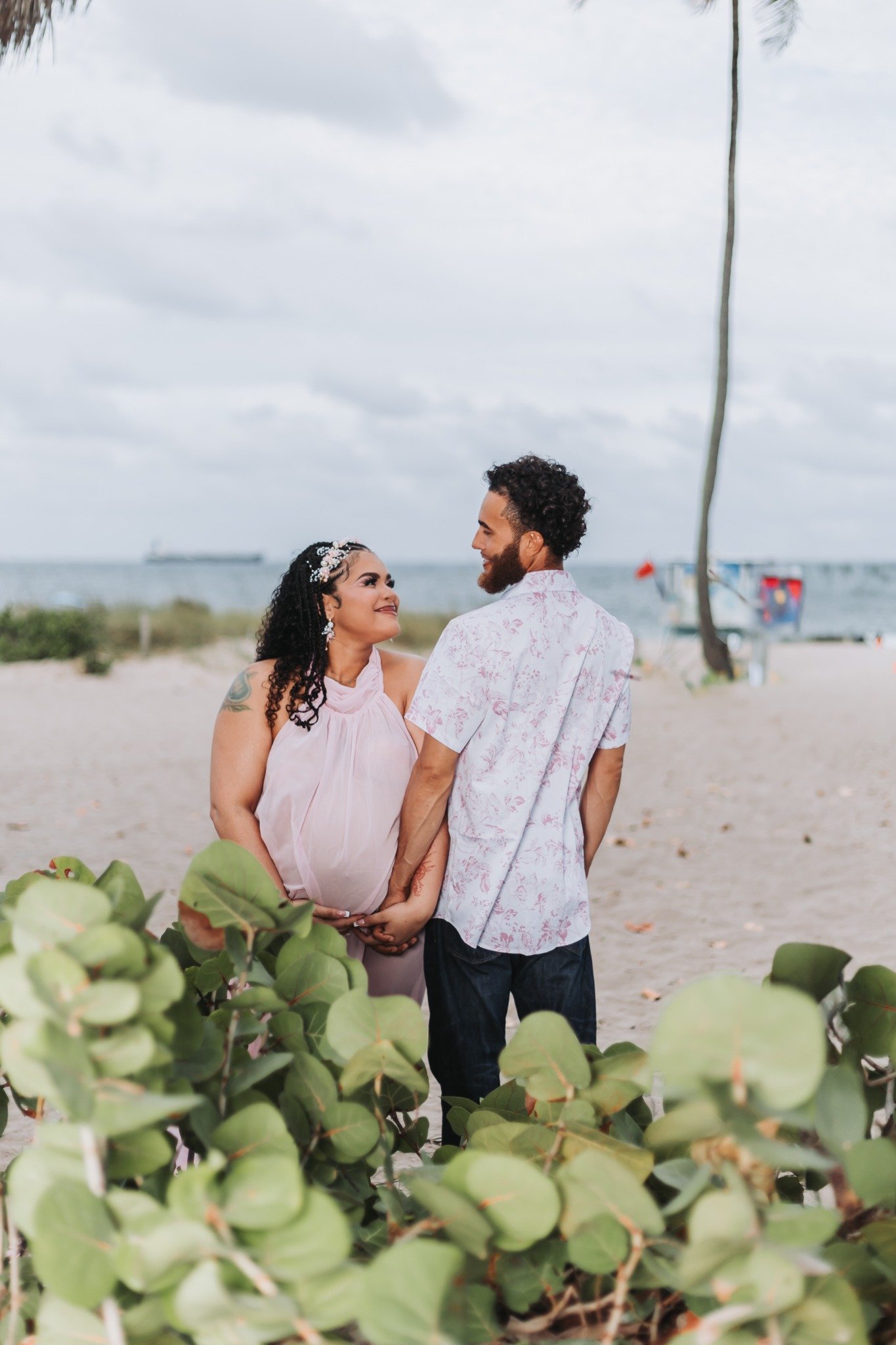 A couple stands on the beach, holding hands and gazing into each other's eyes. The woman has dark, curly hair and tattoos, while the man has a beard and curly hair. There are green leaves in the foreground, and the ocean with a ship and a boat in the