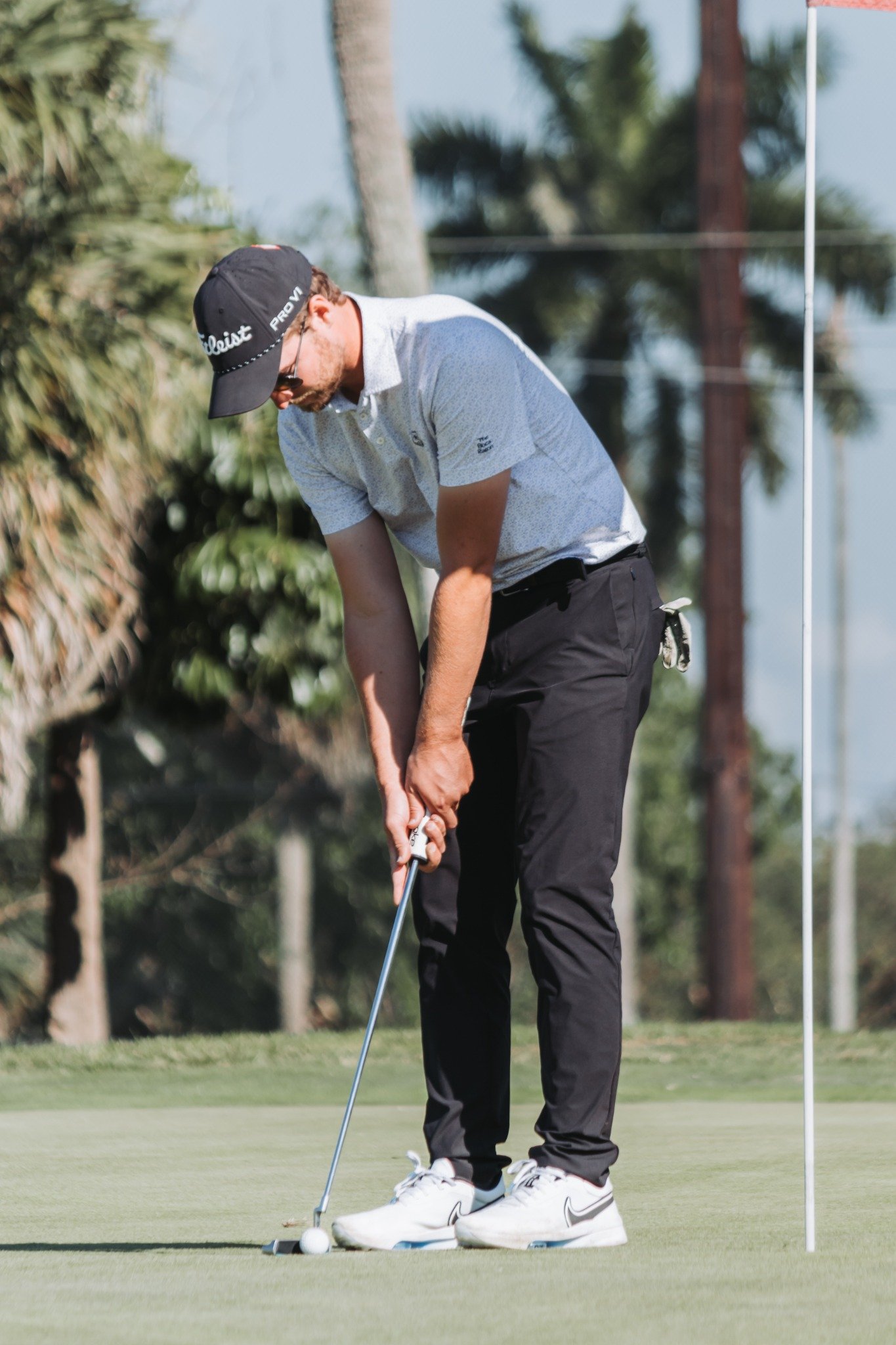 A man putting on a golf glove before putting on the green, with a golf ball and golf hole flag nearby, in a sunny outdoor golf course setting.