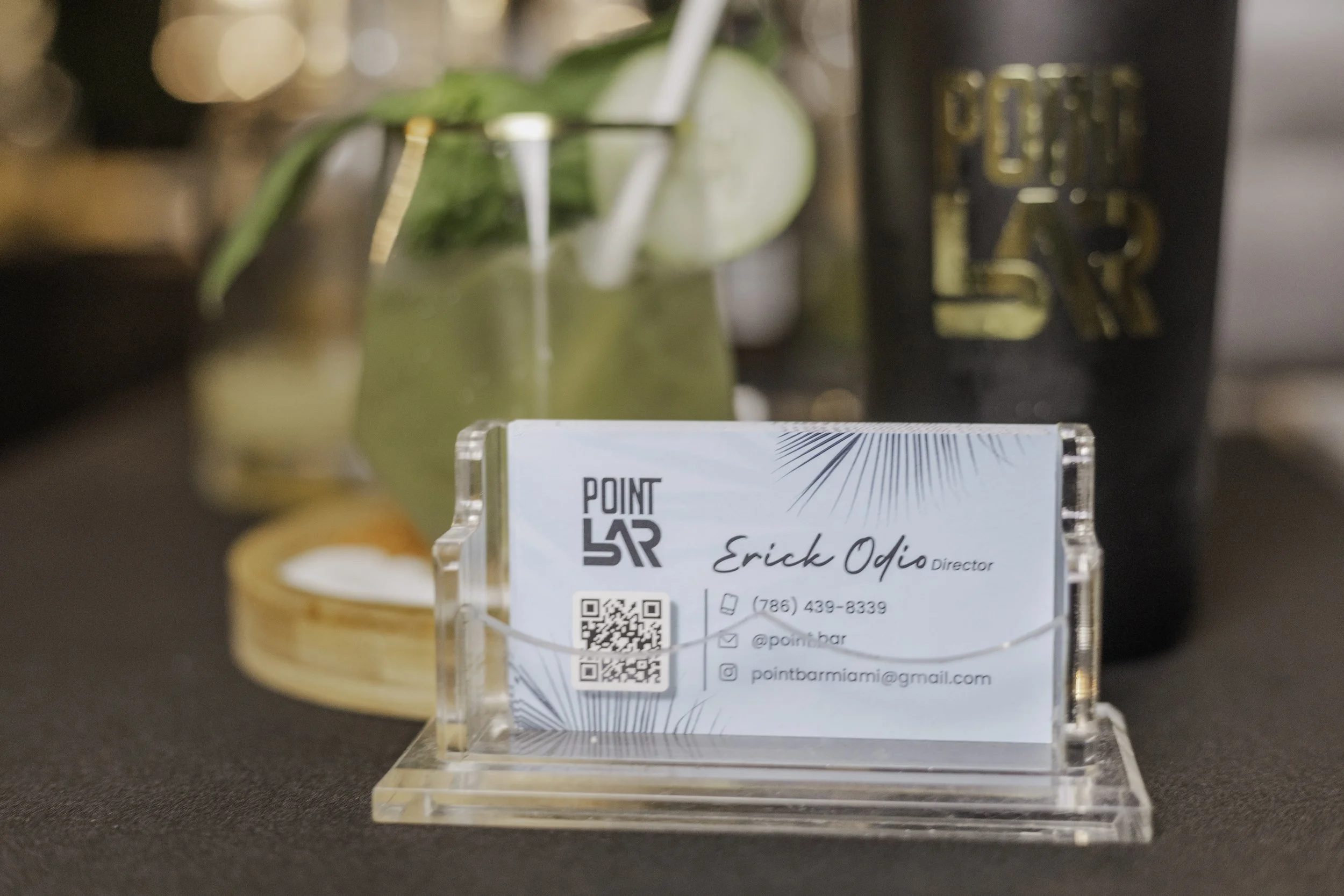 Business card for Point Bar Miami with contact information and QR code, placed on a clear acrylic stand, in front of a mojito cocktail with mint, lime, and straw in the background.