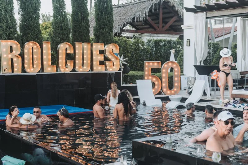 People swimming and socializing in a pool during a celebration at a house, with large illuminated letters spelling 'ROLQUES' and a number '50' in the background, indicating a 50th birthday party.
