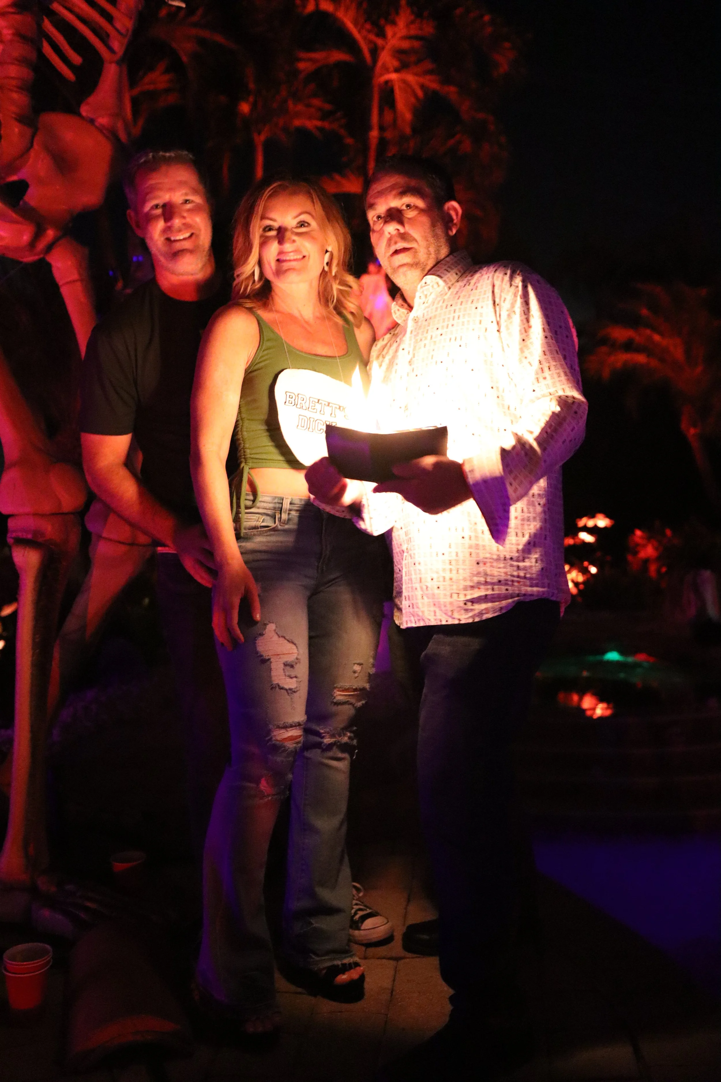 Three people standing close together outdoors at night, with palm trees and dark sky in the background. The woman in the middle is wearing a green top with the words 'Brett's Dig' on it, and the man on the right is holding a lit candle or lantern. Th