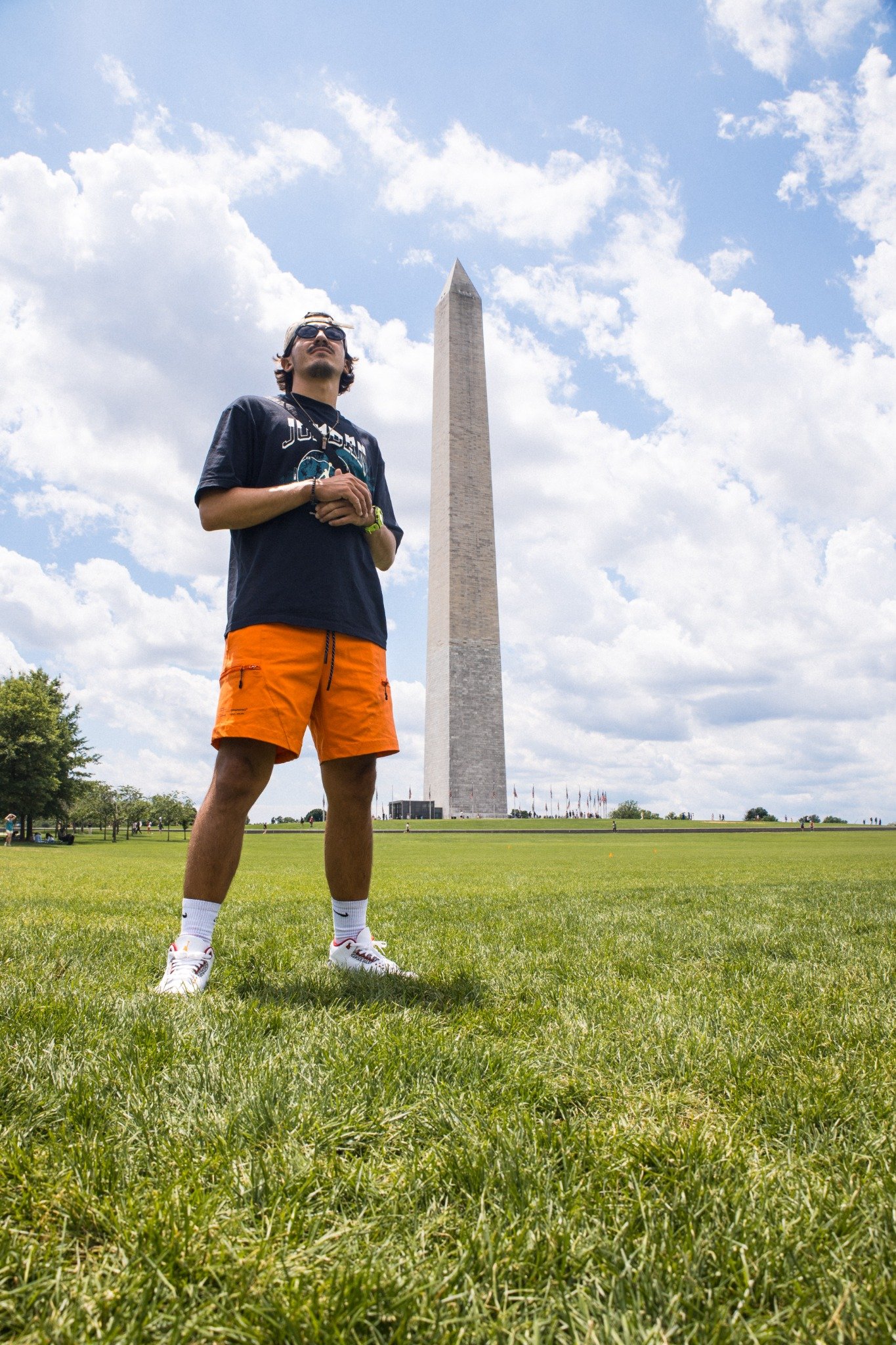 A man wearing sunglasses, a black t-shirt, orange shorts, and white sneakers standing on a grassy field with the Washington Monument in the background and a partly cloudy sky above.