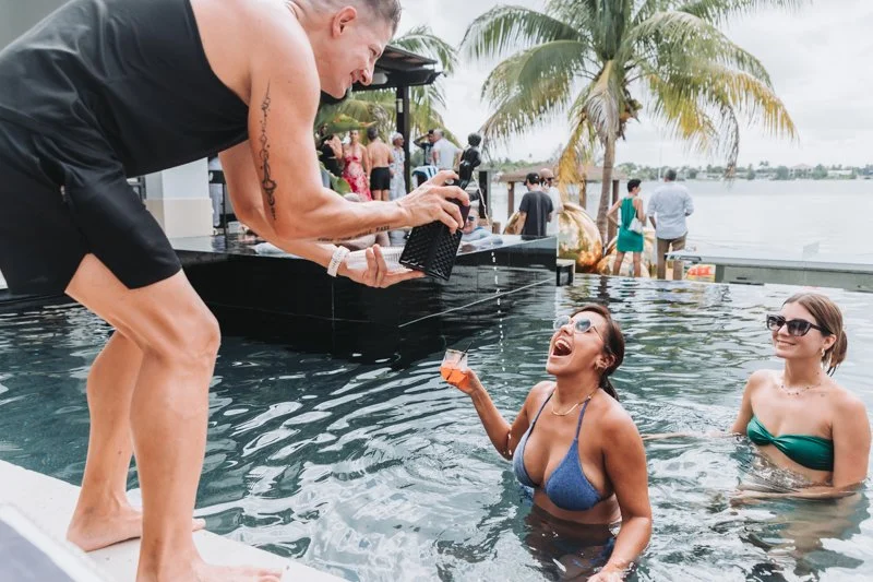 Two women in swimsuits enjoying a pool party near a body of water with palm trees. One woman is receiving a drink from a man on the poolside, and the other woman is standing nearby wearing sunglasses.