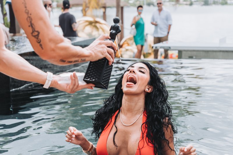 A woman in an orange swimsuit is being baptized in a body of water, with a person pouring water over her head, while others watch in the background.