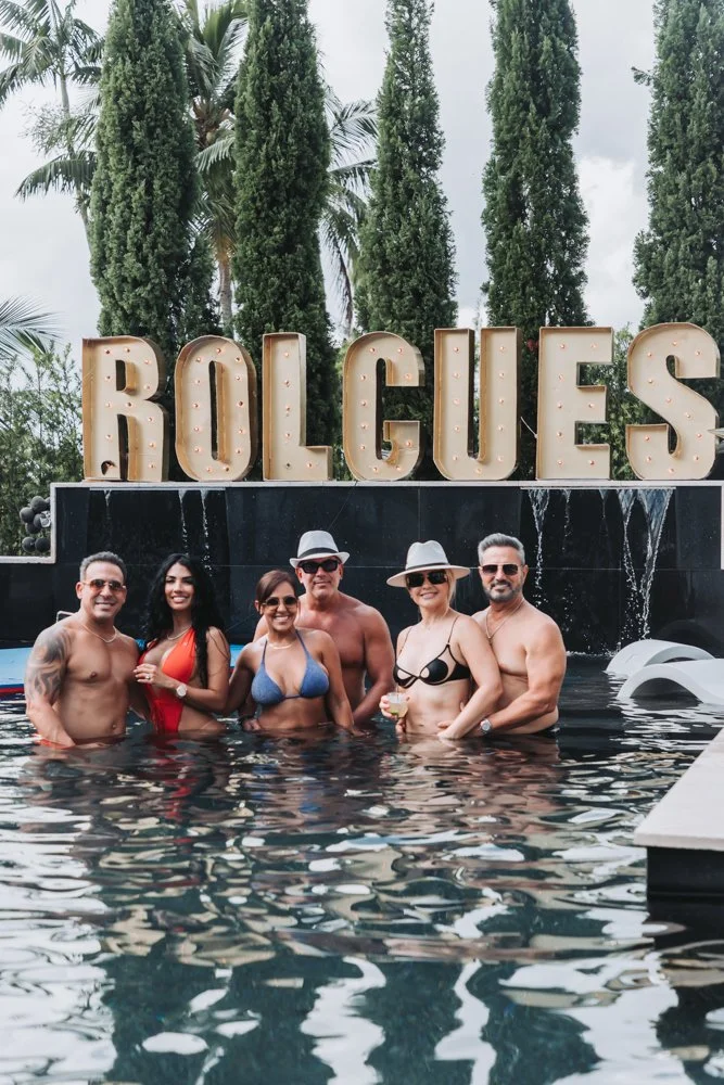Group of six people in a pool with large illuminated spelling sign that reads 'ROLGUES' behind them, surrounded by tall trees and cloudy sky.