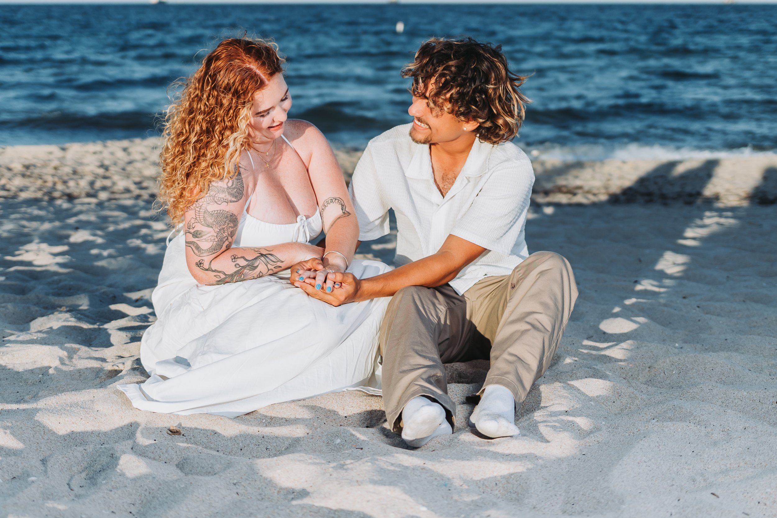 A couple sitting on the sandy beach near the ocean, holding hands and smiling at each other.