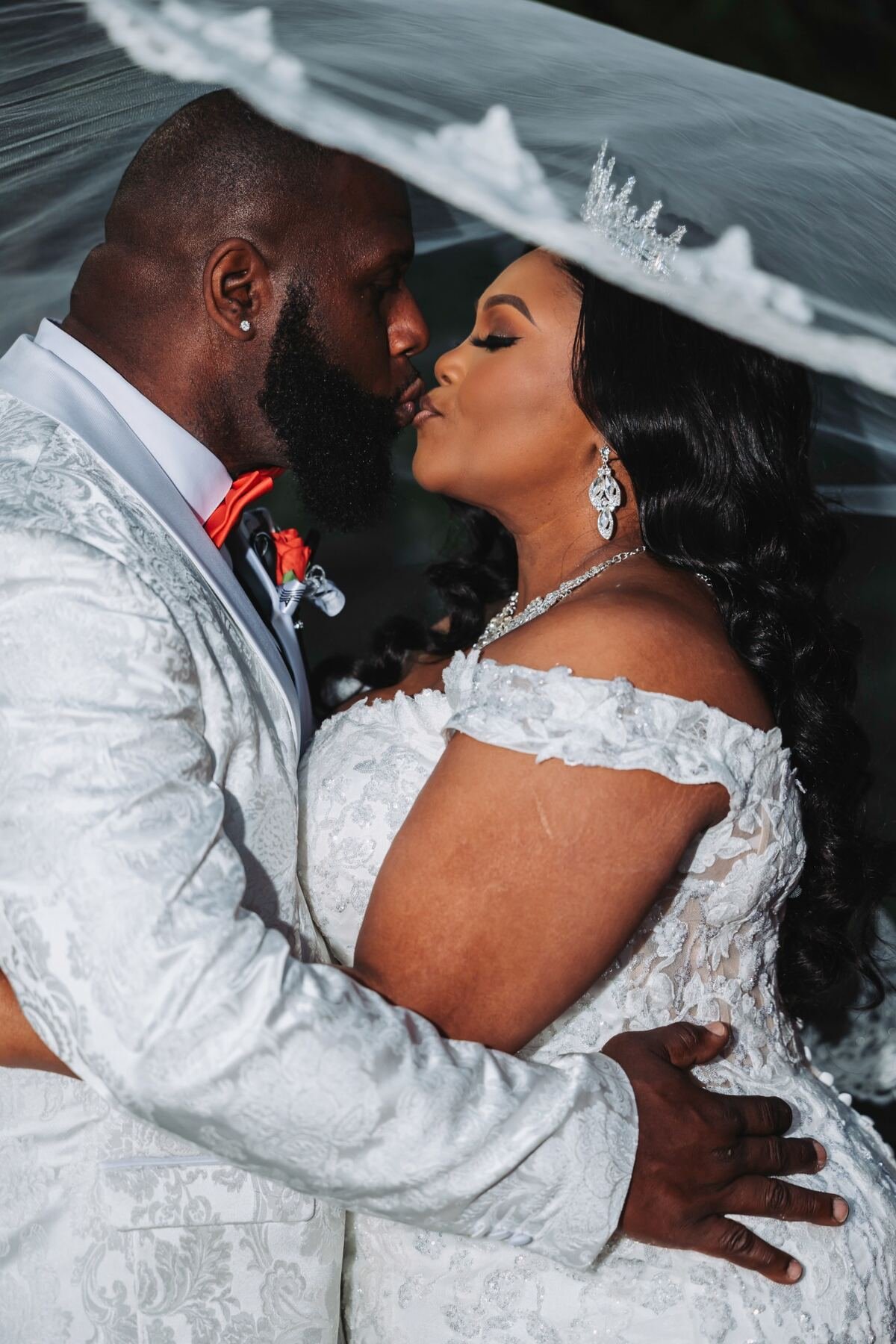 A bride and groom share a kiss under an umbrella, both dressed in white wedding attire, with the bride wearing diamond earrings and a tiara, and the groom dressed in a white suit with a red bow tie.