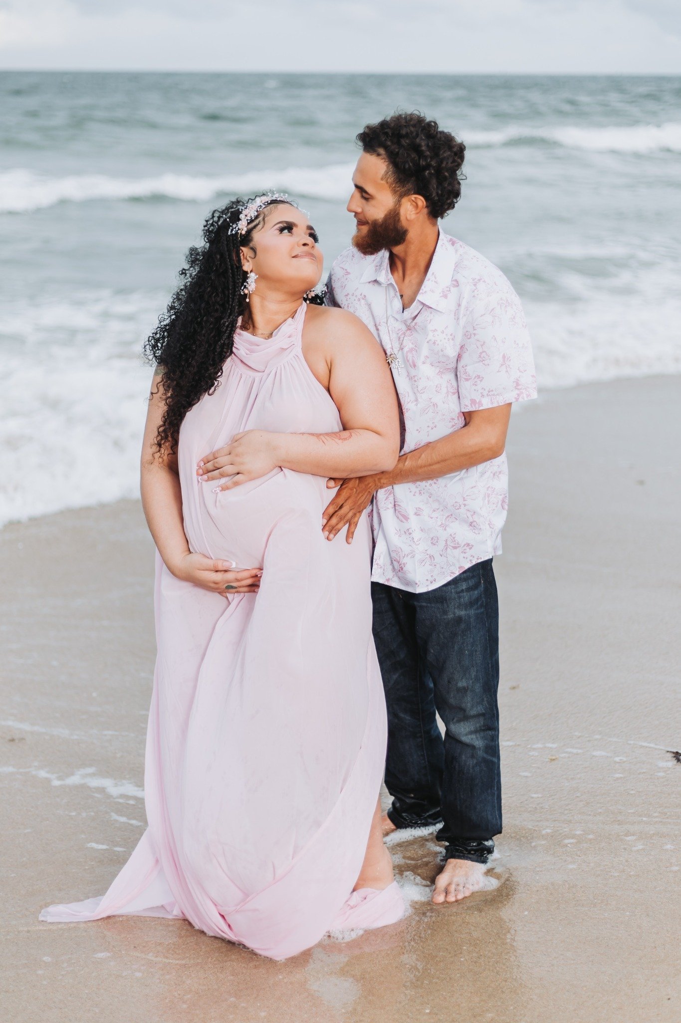A couple standing in the ocean on a beach. The woman is pregnant, wearing a pink dress and a floral headband, looking affectionately at the man. The man has curly hair and a beard, wearing a patterned shirt and jeans, smiling at her. They are barefoo