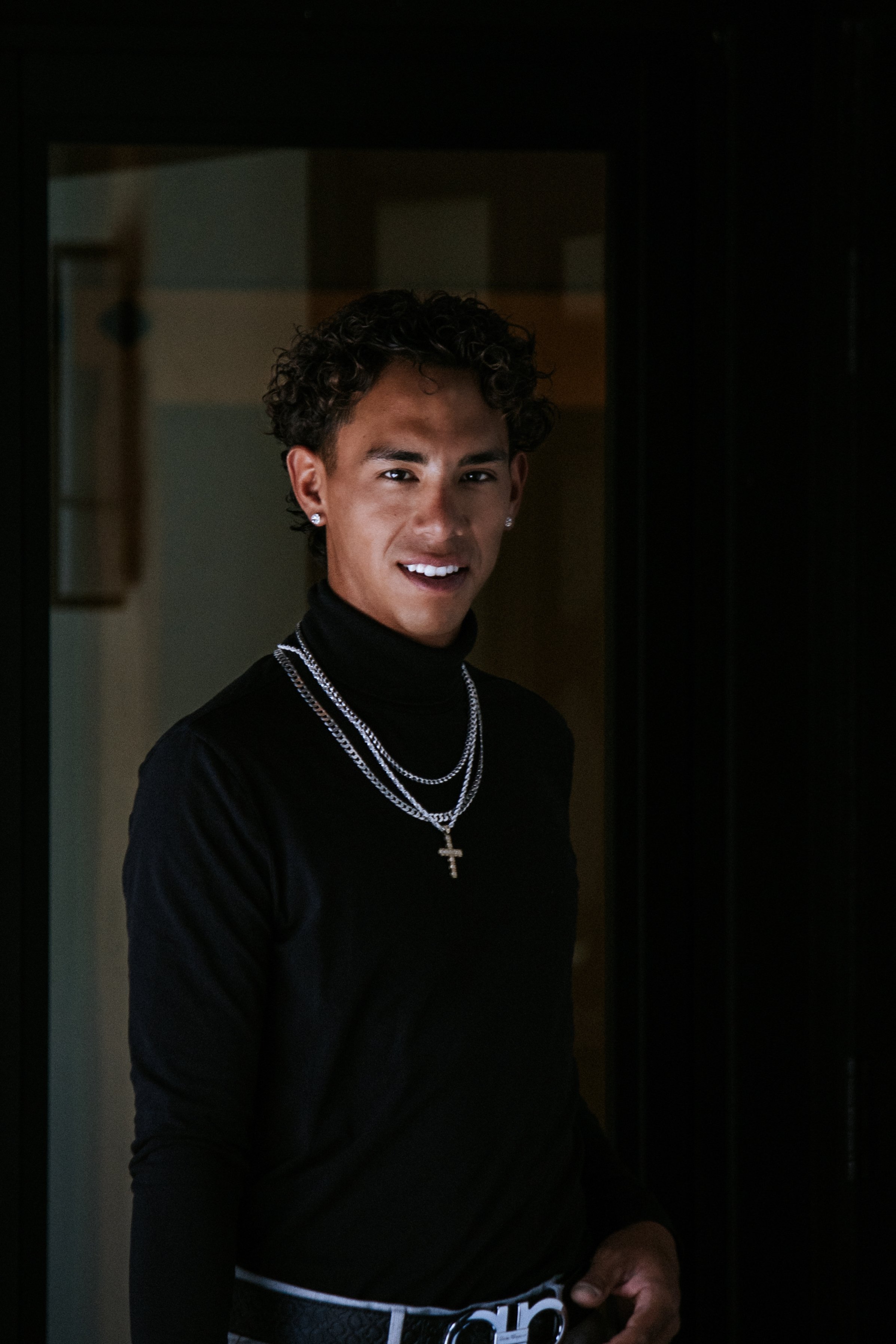 Young man with curly hair and earrings wearing a black turtleneck and layered silver necklaces, standing in a doorway, smiling at the camera.