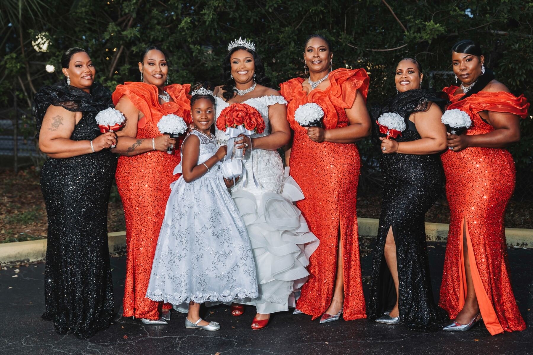 Group of women and a young girl dressed in formal attire, standing outdoors at night with greenery in the background. The women are wearing matching black and red dresses with sequins, and the girl is in a silver gown. The woman in the center is wear
