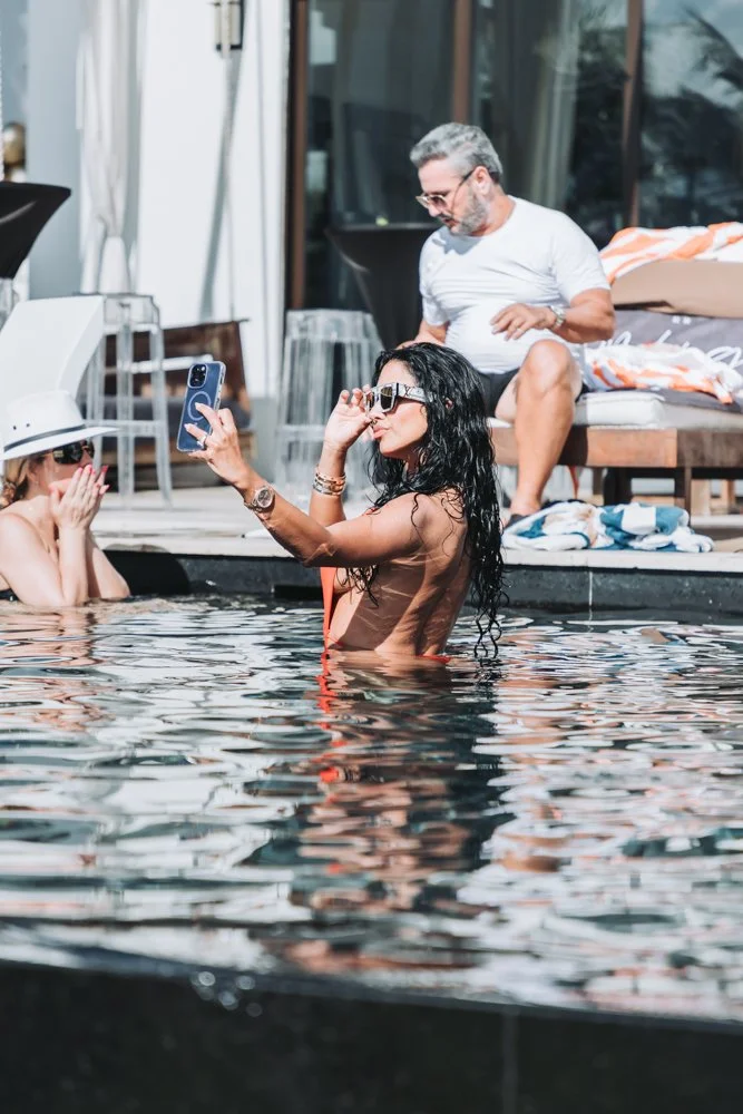 A woman with long curly black hair and sunglasses taking a selfie in a swimming pool with two other women, one wearing a white hat and sunglasses, and a man with gray hair sitting on the poolside behind them.