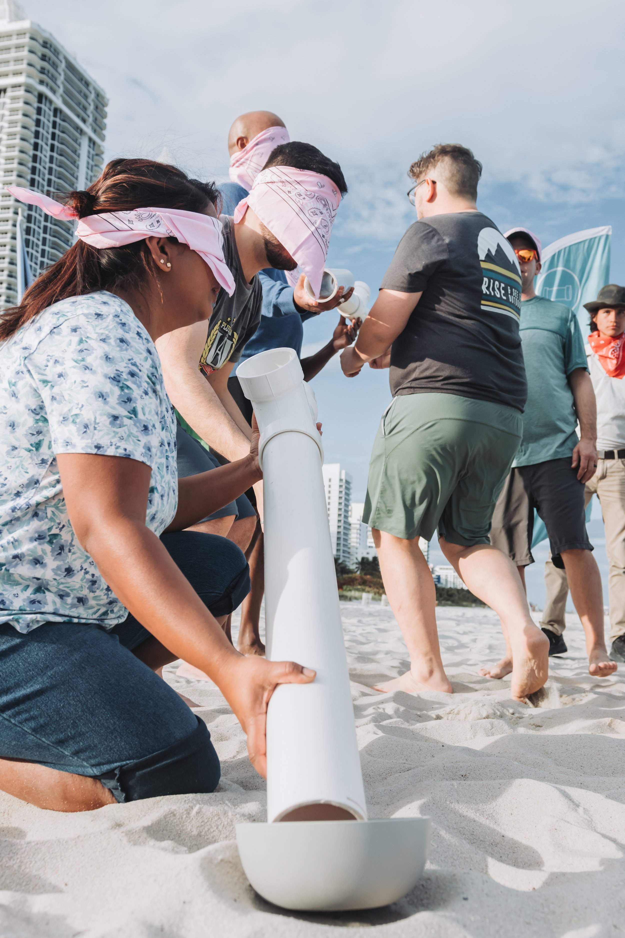 Group of people working on a pipe in the sand on a beach, with tall buildings in the background and cloudy sky.