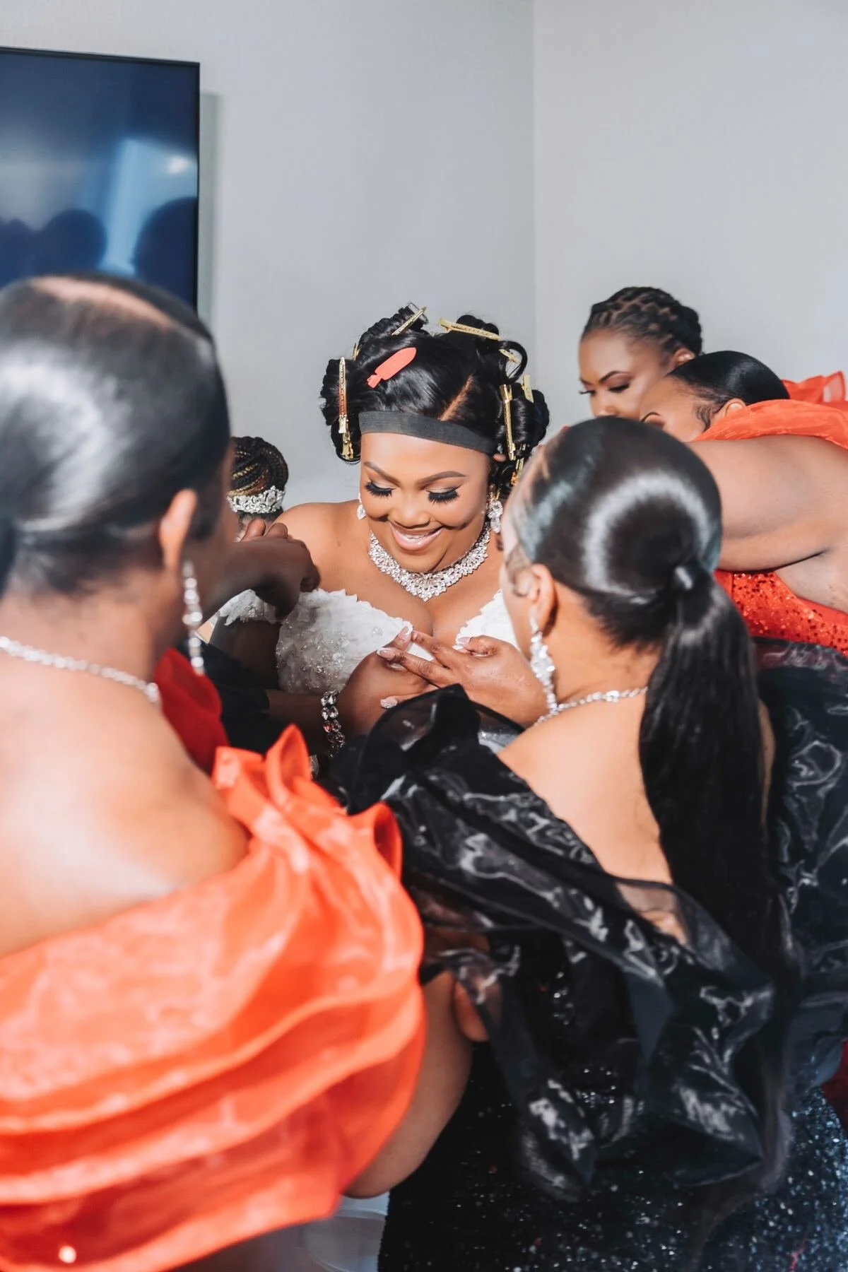 Bride surrounded by women helping her get ready, wearing a wedding dress and jewelry, with hair rollers and clips in her hair.